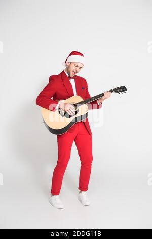 Excited man in suit and red santa hat playing acoustic guitar on grey background Stock Photo