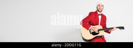 Cheerful man in santa hat playing acoustic guitar on grey background, banner Stock Photo