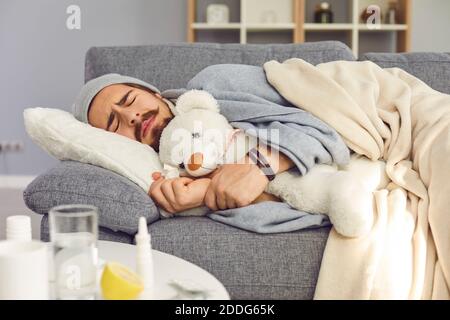 Young unhappy man in warm clothes sleeping under blanket with toy and feeling cold Stock Photo
