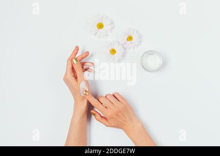 Top view female hands applying cosmetic cream to skin next to chamomile flowers on white background. Stock Photo