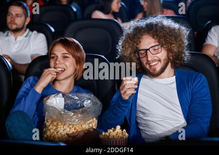 Front view of happy caucasian couple enjoying comedy in cinema together. Young emotional caucasian woman watching film, eating and sharing popcorn from big bag with boyfriend in movie theater hall. Stock Photo