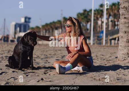 Side view of young female sitting on sandy shore with adorable black Labrador Retriever while enjoying sunset together in summer Stock Photo