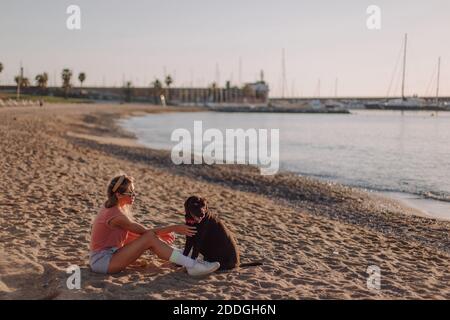 Side view of young female sitting on sandy shore with adorable black Labrador Retriever while enjoying sunset together in summer Stock Photo