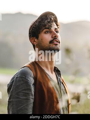 Dreamy young curly haired bearded ethnic male with piercing in ear wearing stylish clothes and cap looking away while standing on green meadow against Stock Photo
