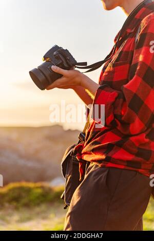 Crop of side view of unrecognizable female traveler with professional photo camera standing on hill in mountainous area Stock Photo