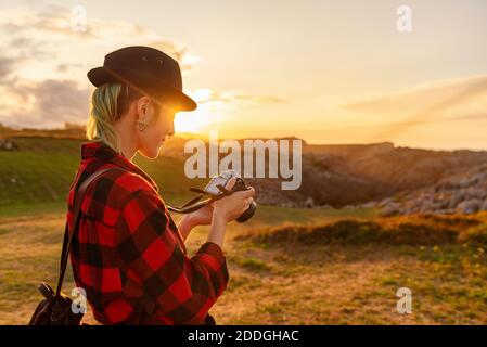 Side view of androgynous female traveler with professional photo camera standing on hill in mountainous area and looking through taken pictures Stock Photo
