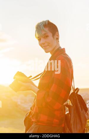 Side view of androgynous female traveler with professional photo camera standing on hill in mountainous area Stock Photo