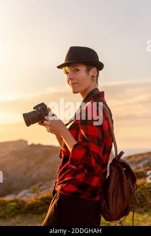 Side view of androgynous female traveler with professional photo camera standing on hill in mountainous area Stock Photo