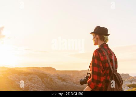 Side view of androgynous female traveler with professional photo camera standing on hill in mountainous area Stock Photo