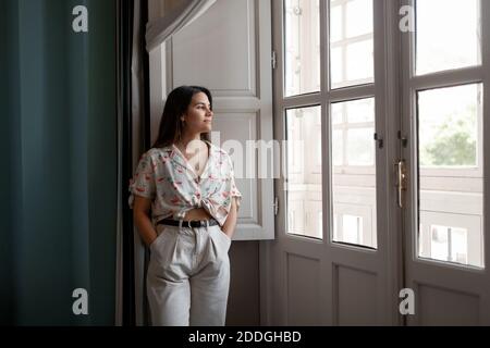 Young carefree female standing with hands in pockets and looking out of window Stock Photo