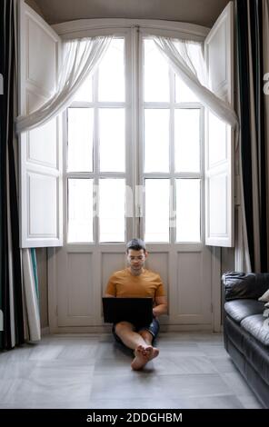Focused male freelancer in casual outfit sitting on floor near window and typing on laptop while working remotely from home Stock Photo