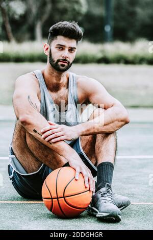 Full body of serious muscular young bearded basketball player in sportswear sitting near ball on street playground and looking at camera Stock Photo