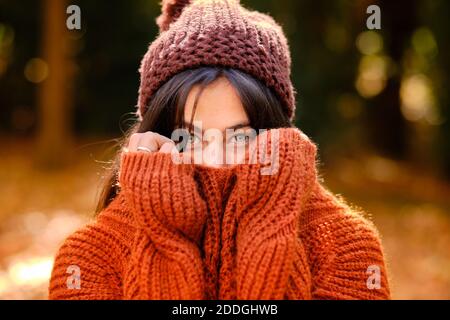 Young female in knitted hat covering face with warm sweater while standing in forest in cold autumn day Stock Photo