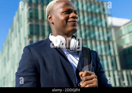 Low angle of stylish African American male entrepreneur with backpack and headphones walking along street in downtown while commuting to work and look Stock Photo