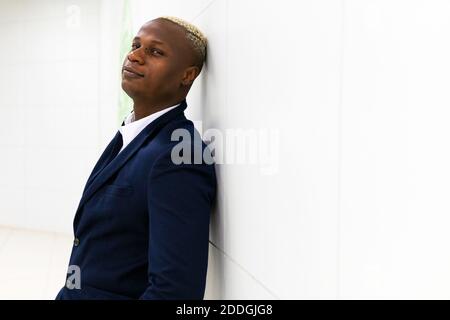 Side view of confident African American male entrepreneur wearing stylish formal suit leaning on wall while standing in white corridor and looking at Stock Photo