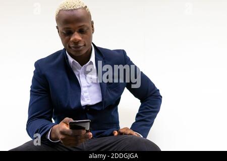 Smiling African American businessman sitting near white wall and messaging on smartphone Stock Photo