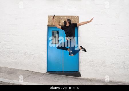 Side view of carefree male in protective mask jumping cheerfully above ground and taking selfie on smartphone while having fun in city Stock Photo