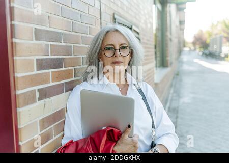 Confident middle aged female entrepreneur in smart casual style standing with laptop along street and looking at camera Stock Photo