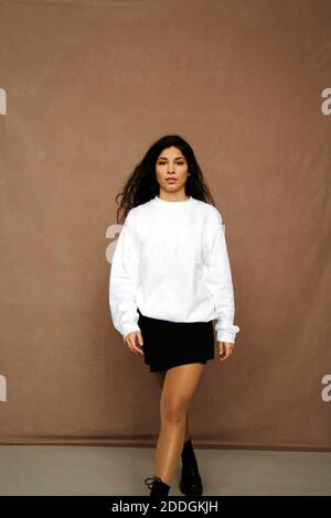 Calm ethnic female in trendy outfit walking on brown background and looking at camera in studio Stock Photo