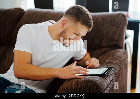 Delighted young male relaxing on couch and surfing Internet on tablet while enjoying weekend at home Stock Photo