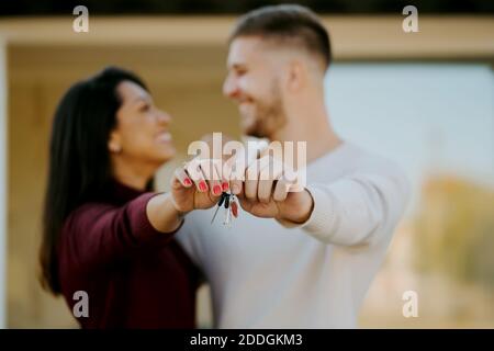 Side view of cheerful couple with keys hugging and looking at camera on background of new house Stock Photo