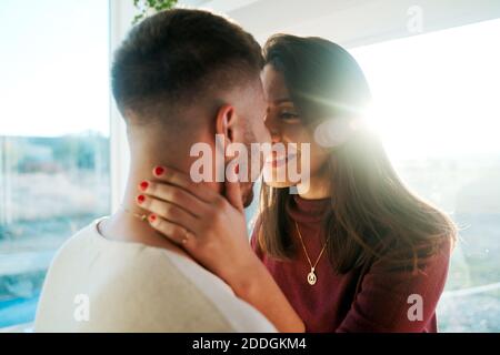 Serene Woman sitting on counter in kitchen and hugging with tender man in romantic bright light Stock Photo
