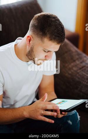 Delighted young male relaxing on couch and surfing Internet on tablet while enjoying weekend at home Stock Photo