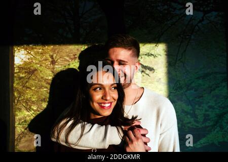 Cheerful man embracing smiling Woman from back while standing in apartment lit by sunlight Stock Photo