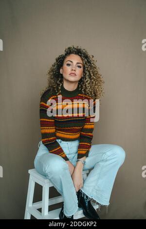 Delighted female with Afro hairstyle sitting on wooden chair in studio and looking at camera Stock Photo