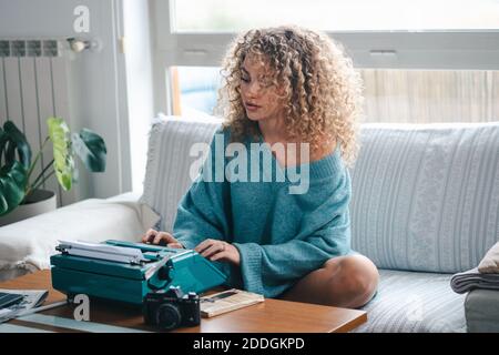 Focused female author sitting on sofa and using vintage typewriter while creating interesting story Stock Photo