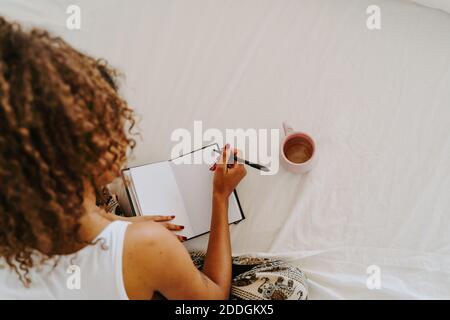 From above back view of unrecognizable female sitting on bed with cup of coffee and taking notes in diary while making plans for week at home Stock Photo