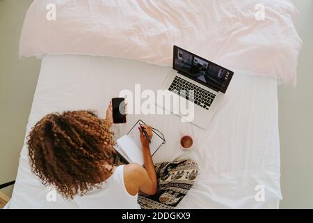 From above crop unrecognizable focused young African American female freelancer in casual clothes making notes in planner with smartphone while sittin