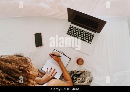 From above crop unrecognizable focused young African American female freelancer in casual clothes making notes in planner while sitting on bed with la