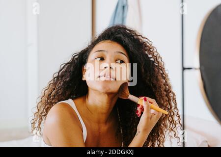 Young curly haired ethnic female applying blush from palette on cheek with cosmetic brush during daily makeup procedure at home Stock Photo