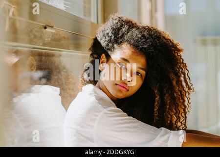 Side view of young African American female with gorgeous long curly hair looking at camera while sitting near window in sunlight Stock Photo