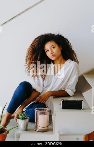 Attractive young barefoot ethnic female with long afro hairstyle dressed in casual white blouse and jeans sitting on stairs near potted plants and can Stock Photo