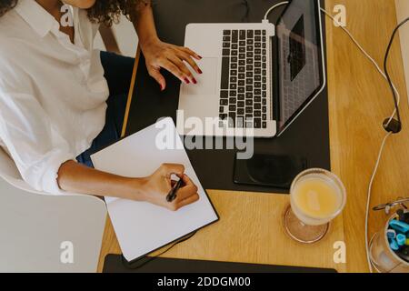 From above cropped unrecognizable female remote employee in casual outfit sitting at table with laptop and writing in planner while working in home of Stock Photo