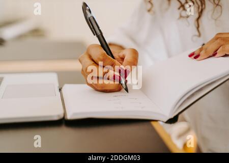 Crop unrecognizable female entrepreneur sitting at table with laptop and writing plan for business project in notebook Stock Photo