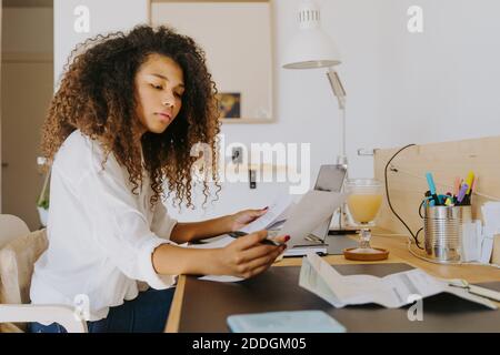 Side view of female bookkeeper sitting at table in home office and analyzing financial reports and paper documents during remote work Stock Photo