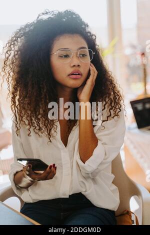 Serious African American female entrepreneur with erabuds sitting at table in home office and reading messages on cellphone during distant work Stock Photo