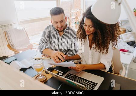 High angle of content multiracial couple of entrepreneurs sitting at table and reading paper documents while working from home Stock Photo