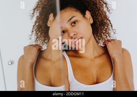 Calm young ethnic female with Afro hairstyle looking at camera while standing near mirror in morning at home Stock Photo