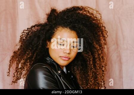 Young African American female with curly hair wearing trendy leather jacket standing on pink background looking down Stock Photo