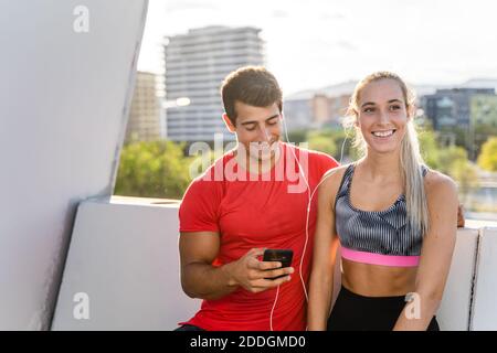 Young slim Woman sportswear and strong muscular man listening to music on earphones on smartphone after fitness workout together in city Stock Photo