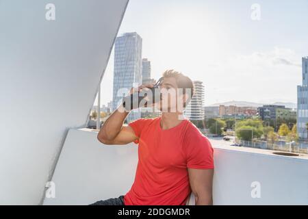 Young muscular male in sportswear drinking water from plastic bottle while resting after intense workout on terrace of modern city building Stock Photo