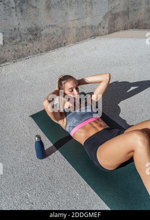 From above of focused female athlete doing abdominal crunches while lying on mat and training in city on sunny day Stock Photo