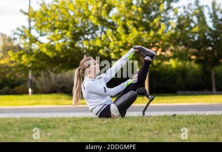 Side view of professional female runner with bionic prosthesis stretching legs during workout while sitting on grass Stock Photo