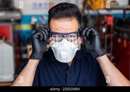 Adult man in latex gloves and respirator putting on protective goggles and looking at camera during work in garage Stock Photo