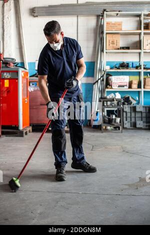 Low angle of man in uniform sweeping floor with broom while working in garage Stock Photo