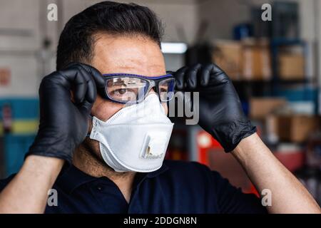Adult man in latex gloves and respirator putting on protective goggles and looking at camera during work in garage Stock Photo